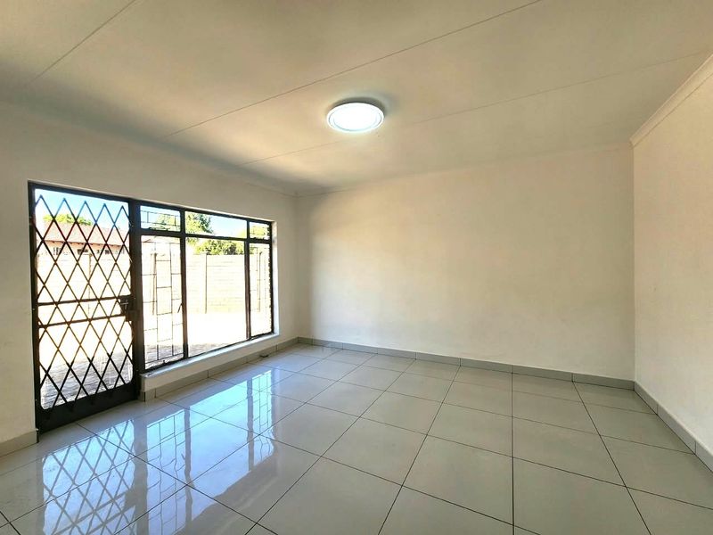 A bright, empty office space featuring polished tiled flooring, a large window with a security gate, and neutral wall finishes.