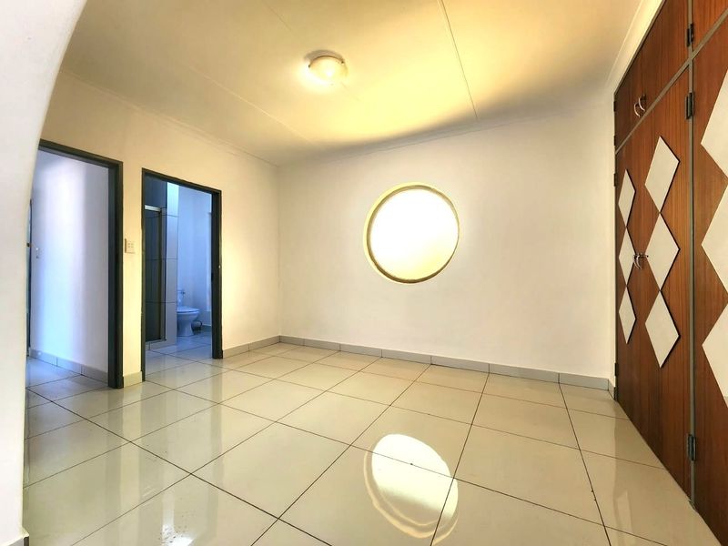 A bright, empty office space featuring tiled flooring, a unique circular window, and built-in wooden cabinetry, with direct access to an adjacent bathroom.