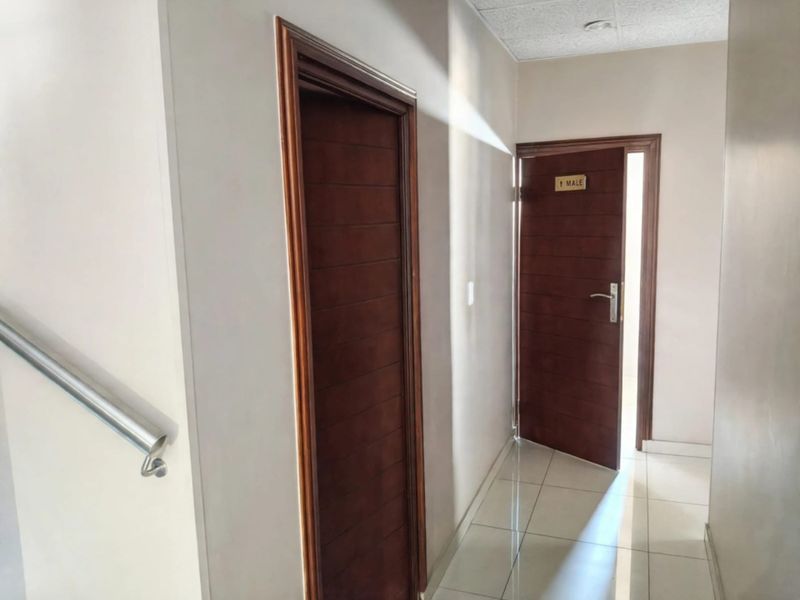 A clean, well-lit office hallway featuring tiled floors, a stainless steel handrail, and wooden doors leading to separate facilities.