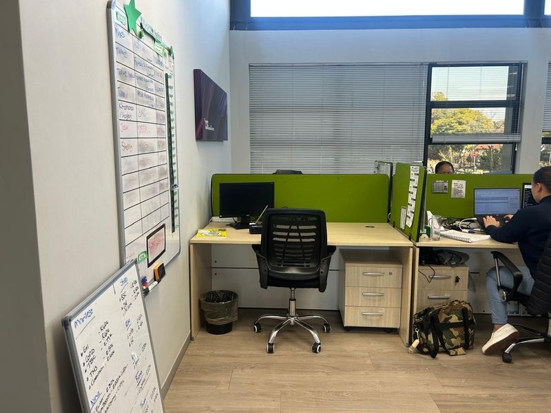 A functional office space featuring laminate flooring, built-in desk workstations with integrated storage, and large windows with venetian blinds for natural light.