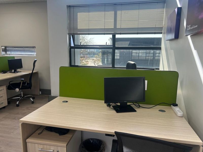 Modern office workspace featuring built-in desks with privacy screens, laminate flooring, and large windows with venetian blinds providing natural light.
