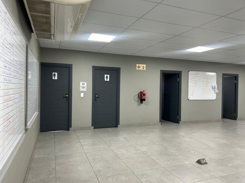 A clean, well-maintained industrial hallway featuring tiled flooring, suspended ceilings with recessed lighting, and multiple access doors to restroom facilities.
