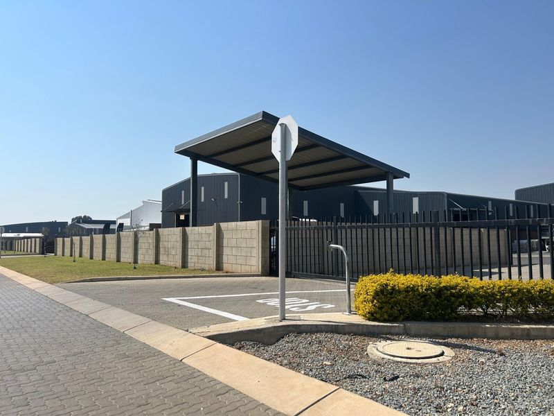 Exterior view of an industrial property featuring a paved parking area, metal carport, and secure perimeter fencing with a concrete block wall.