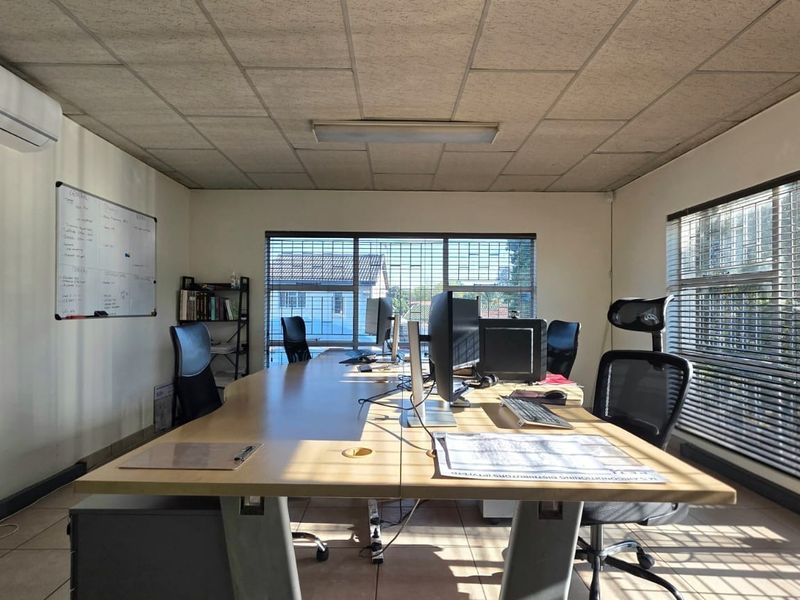 A functional office space featuring tiled flooring, suspended ceiling tiles, split-system air conditioning, and large windows with security bars and venetian blinds.