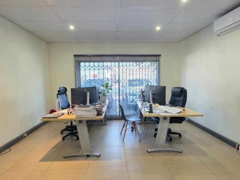 A functional office space featuring tiled flooring, a suspended ceiling with recessed lighting, and a wall-mounted air conditioning unit.
