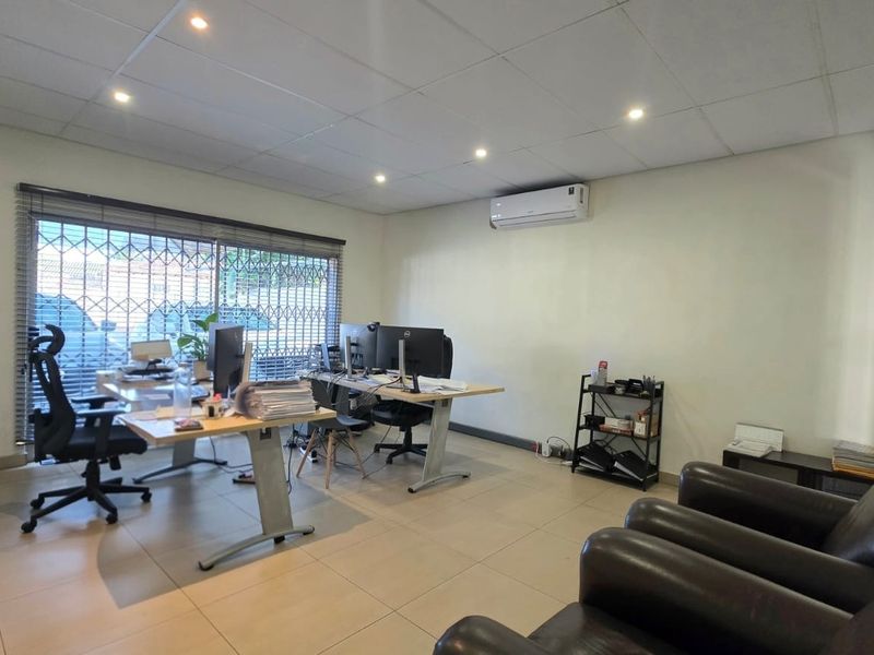 A functional office space featuring tiled flooring, a suspended ceiling with recessed lighting, and a wall-mounted air conditioning unit.