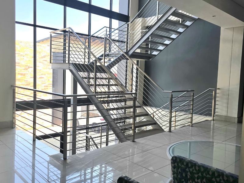 Modern office entrance featuring a striking stainless steel staircase, floor-to-ceiling glass windows, and polished tiled flooring, creating a bright and professional atmosphere.