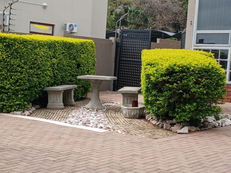 A paved rear exterior area featuring a landscaped garden bed with manicured hedges, stone accents, and secure boundary walling with electric fencing.