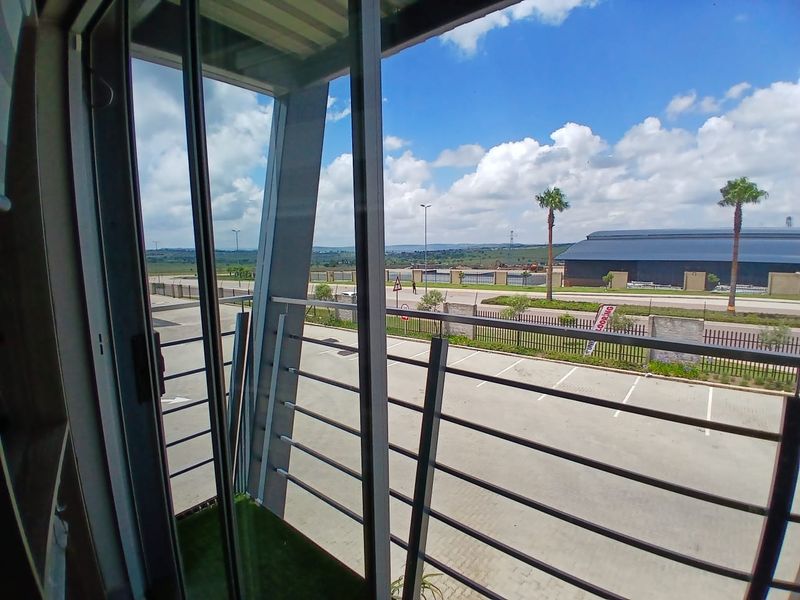 A modern balcony featuring a glass sliding door, metal safety railing, and artificial turf flooring, overlooking a paved parking area and industrial landscape.