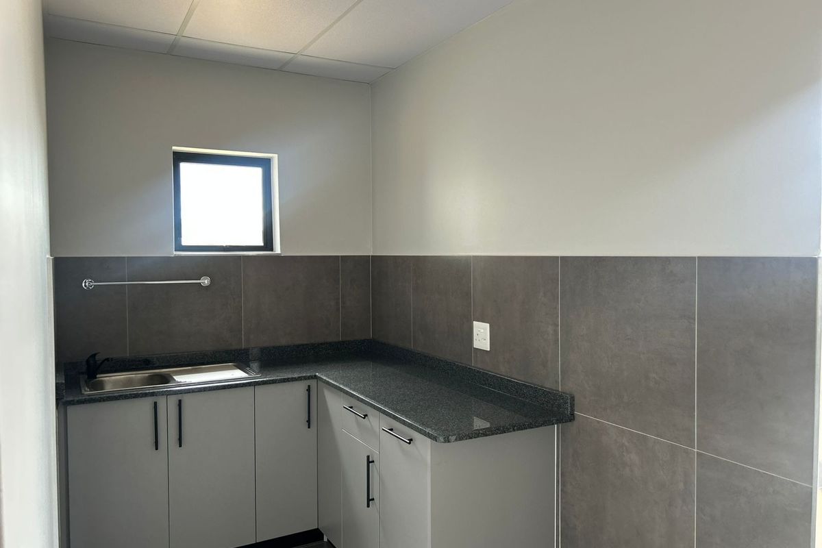 A clean, modern kitchenette featuring grey built-in cabinetry, dark granite countertops, a stainless steel sink, and neutral tiled splashbacks.