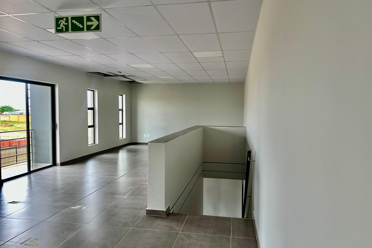 Spacious industrial hallway featuring tiled flooring, a suspended ceiling with an emergency exit sign, and access to a stairwell and exterior balcony.