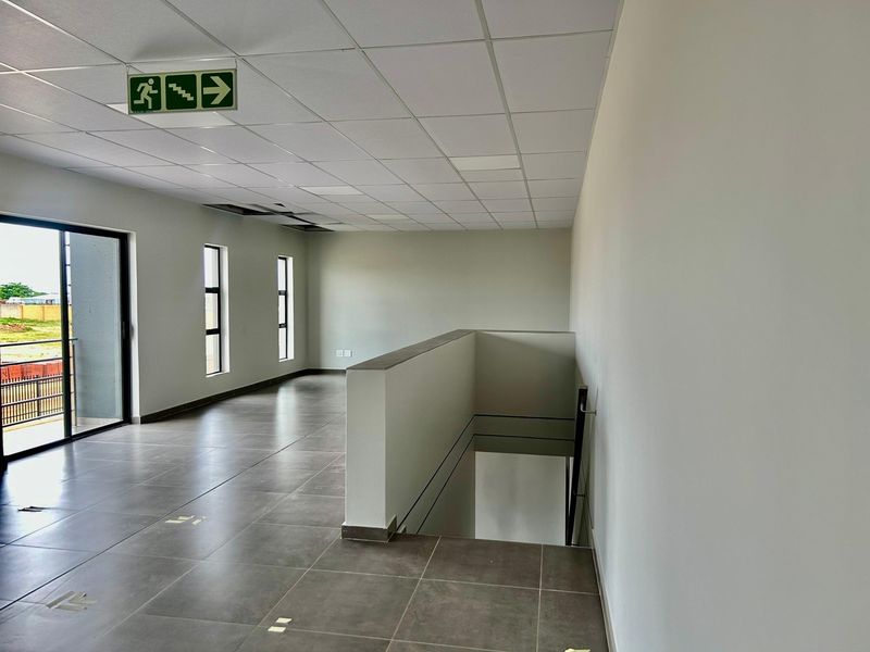 Spacious industrial hallway featuring tiled flooring, a suspended ceiling with an emergency exit sign, and access to a stairwell and exterior balcony.