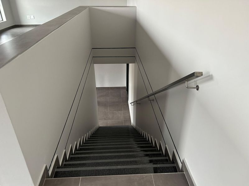 A modern, minimalist staircase featuring dark tiled steps, clean white walls, and a sleek stainless steel handrail leading to a lower level.