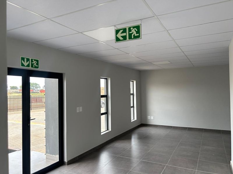 A clean, modern, and empty office space featuring neutral grey walls, large aluminium windows, tiled flooring, and a suspended ceiling with emergency exit signage.