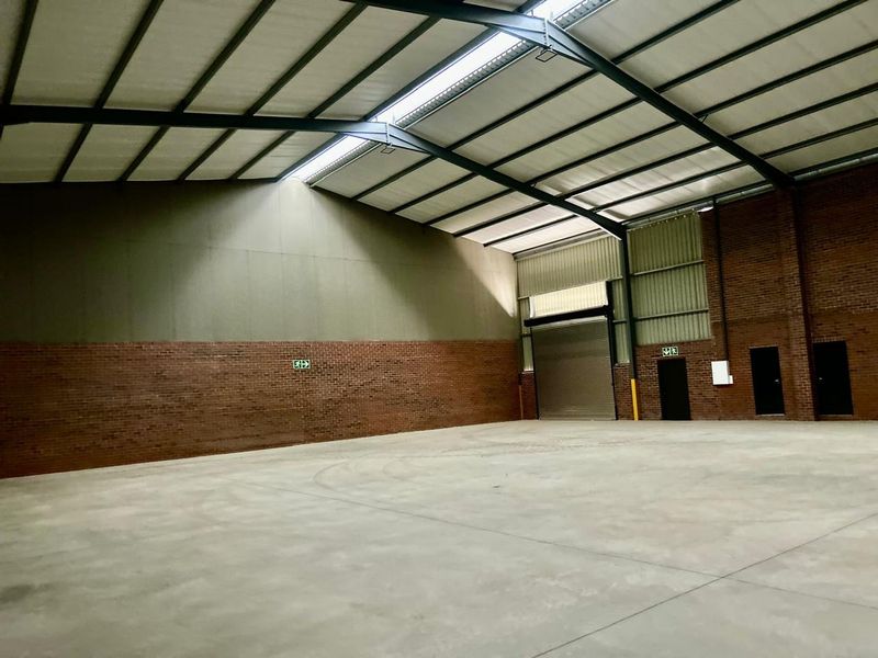 Spacious industrial warehouse unit featuring high ceilings with exposed steel trusses, concrete flooring, brick walls, and a large roller shutter door for easy access.