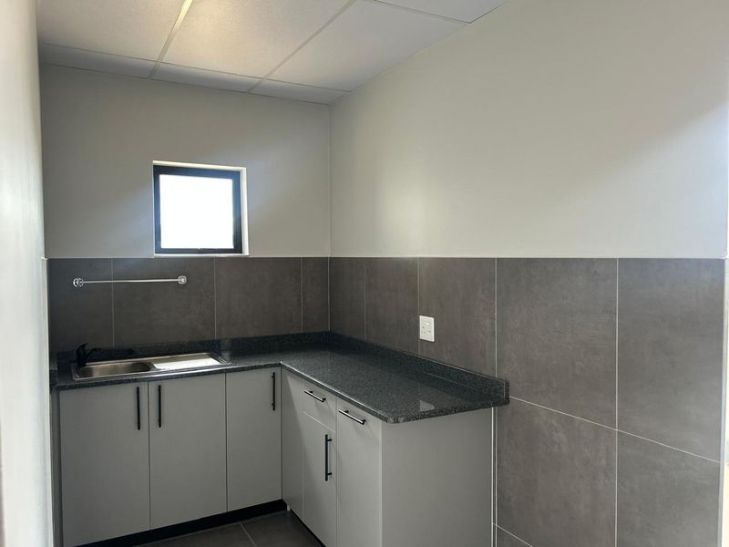 A clean, modern kitchenette featuring grey cabinetry, granite countertops, a stainless steel sink, and neutral tiled wall finishes.