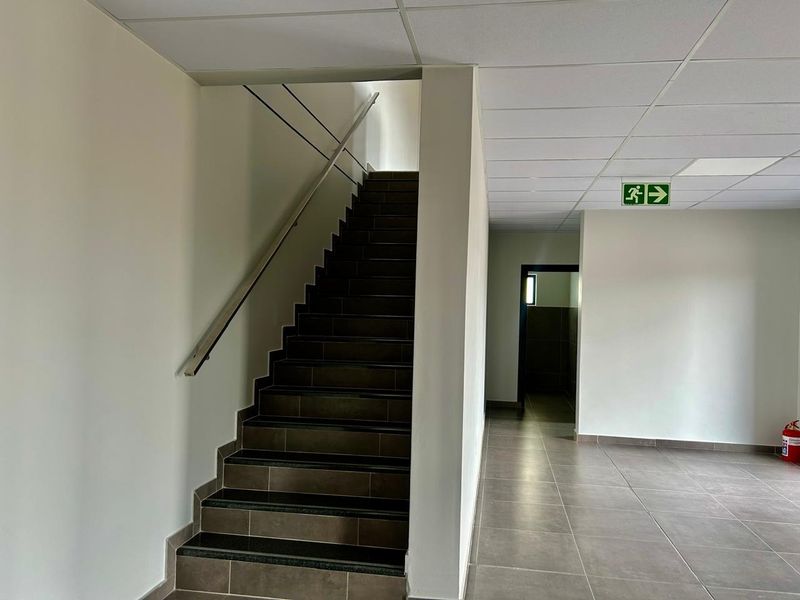 A clean, modern industrial hallway featuring a tiled staircase with a metal handrail, suspended ceiling, and clear emergency exit signage.