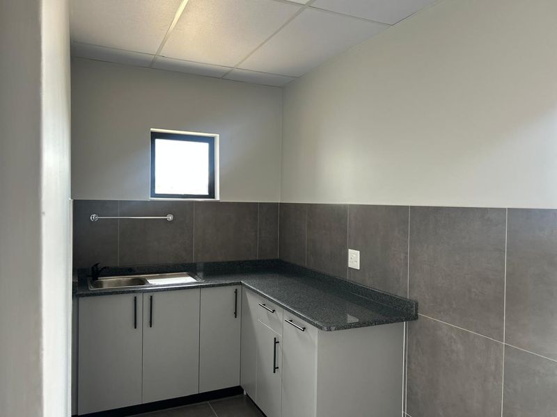 A clean, modern kitchenette featuring grey cabinetry, granite countertops, a stainless steel sink, and neutral tiled backsplash in an industrial setting.