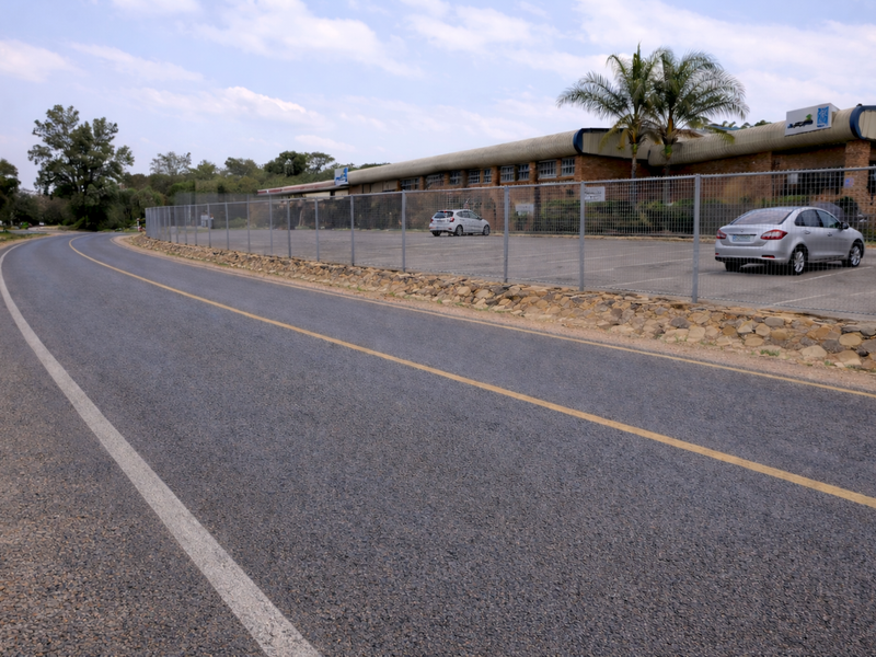Street-level view of the commercial property featuring a paved parking area secured by perimeter fencing and a stone retaining wall.