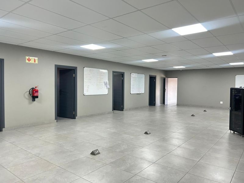 A spacious, modern commercial office area featuring neutral grey walls, tiled flooring, recessed ceiling lighting, and multiple access doors.
