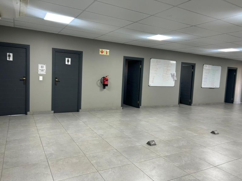 A spacious commercial hallway featuring tiled flooring, recessed ceiling lighting, and multiple access doors leading to various office or restroom facilities.