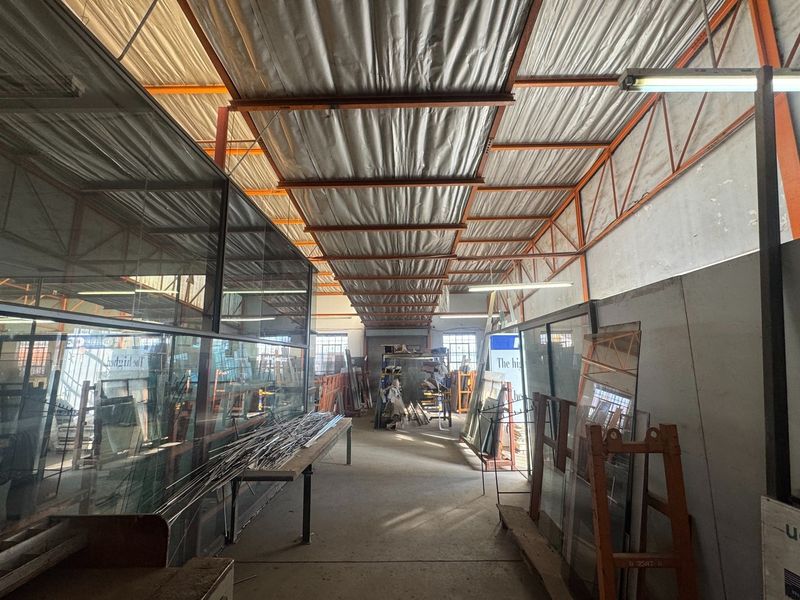 Industrial utility space featuring exposed steel roof trusses, concrete flooring, and a partitioned glass office enclosure, suitable for workshop or storage use.