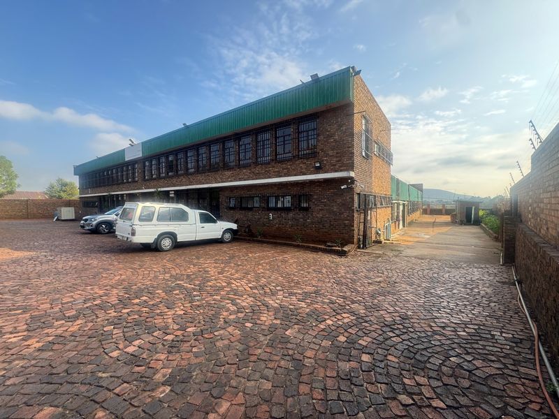 Exterior view of a multi-story industrial brick building featuring a cobblestone driveway, security bars on windows, and perimeter wall with electric fencing.