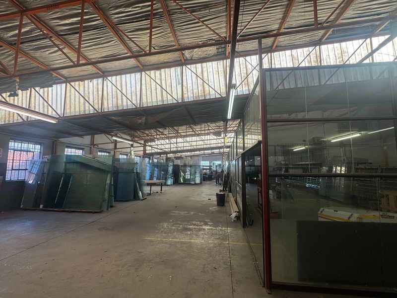 Spacious industrial warehouse area featuring high exposed steel roof trusses, translucent skylights, concrete flooring, and a partitioned glass office enclosure.