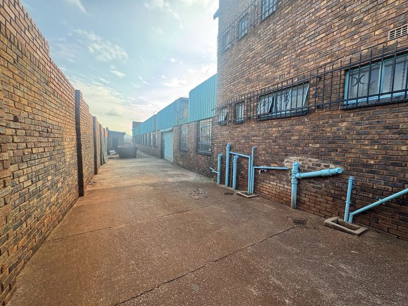 Rear exterior view of an industrial property featuring exposed brick walls, security bars on windows, and external plumbing infrastructure on a concrete paved area.