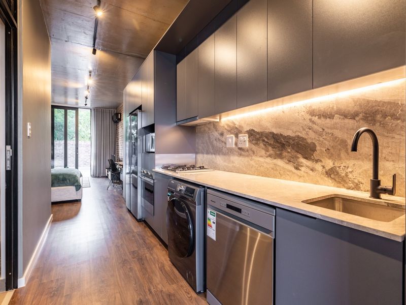 Modern open-plan kitchen featuring sleek dark cabinetry, stone backsplash, integrated appliances, and stylish track lighting, seamlessly flowing into the living area.