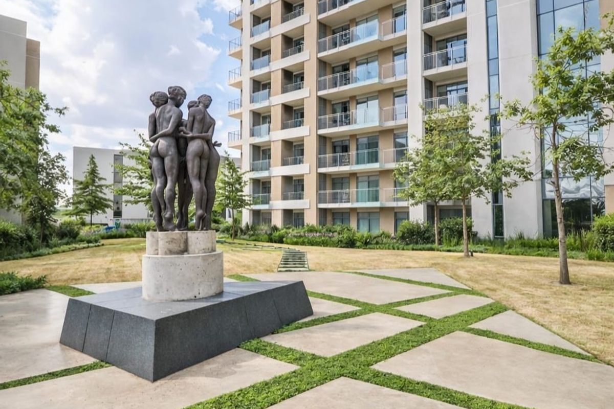 A well-maintained communal garden area featuring a central sculpture, concrete paving, and a view of the modern apartment building facade with balconies.