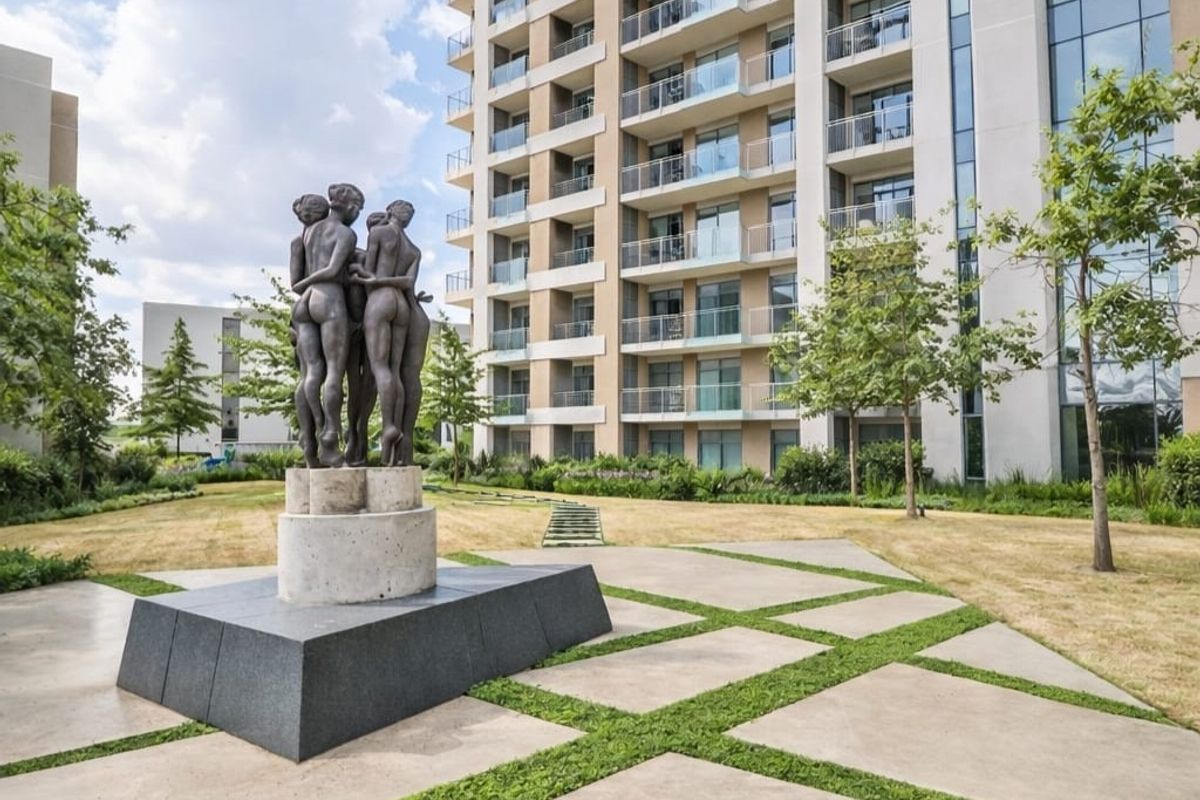 A well-maintained communal garden area featuring a central sculpture, paved pathways, and views of the modern apartment building facade with private balconies.