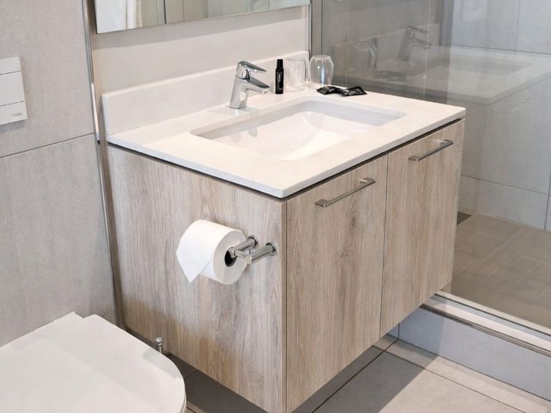 Modern bathroom featuring a wall-mounted wood-finish vanity with integrated basin, wall-hung toilet, and a glass-enclosed shower with neutral tiling.
