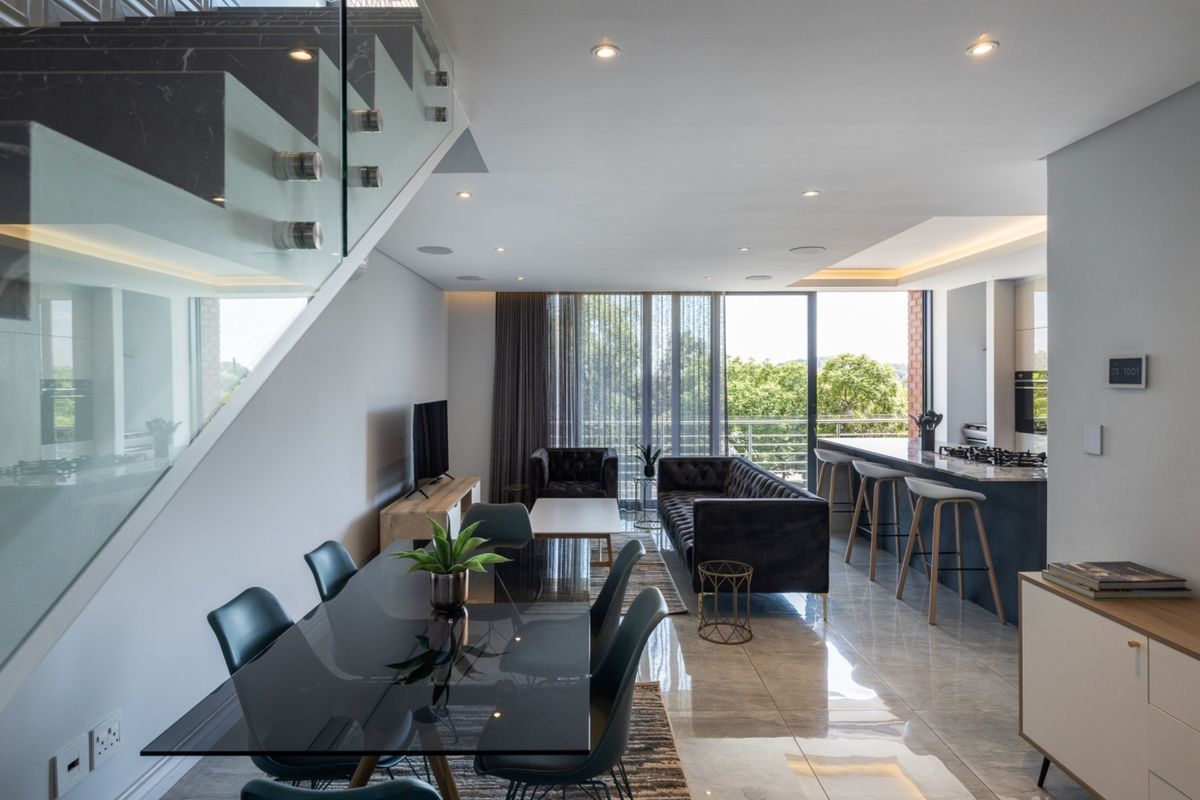 Modern open-plan living area featuring a sleek kitchen with stone island, glass staircase balustrade, and seamless flow to an outdoor balcony.