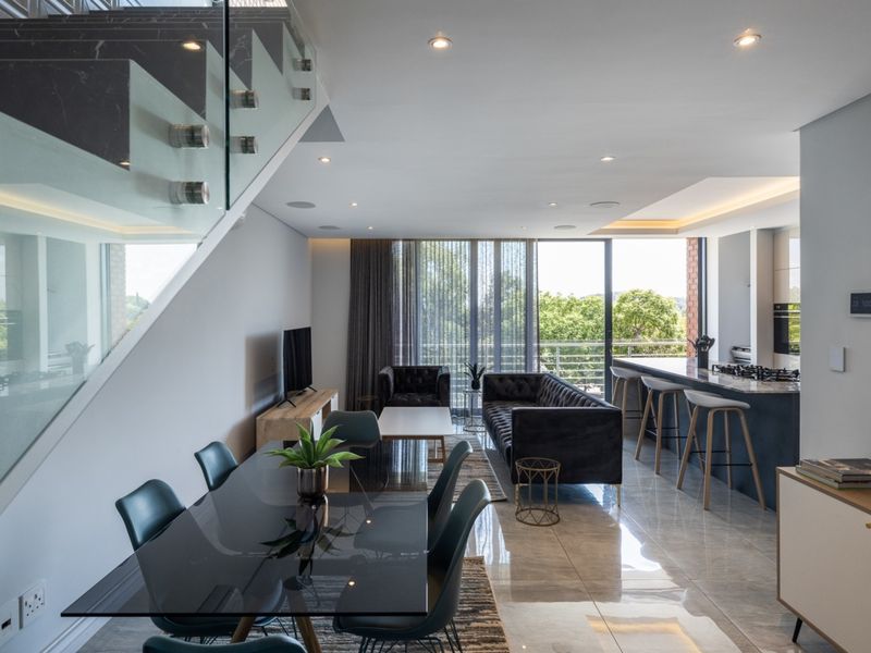 Modern open-plan living area featuring a sleek kitchen with stone island, glass staircase balustrade, and seamless flow to an outdoor balcony.