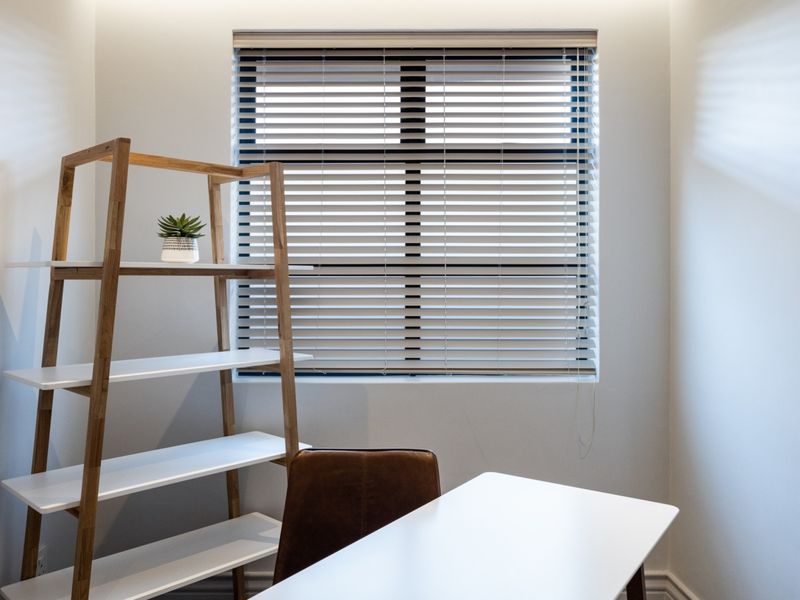 A bright and well-maintained home office space featuring clean white walls and fitted venetian blinds.