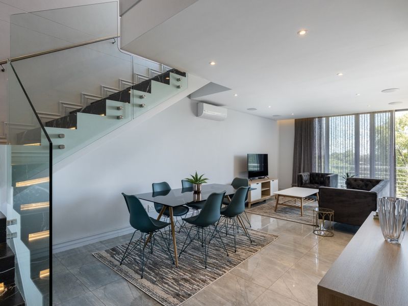 Modern open-plan living and dining area featuring a striking glass-balustrade staircase with marble treads, tiled flooring, and large windows providing natural light.