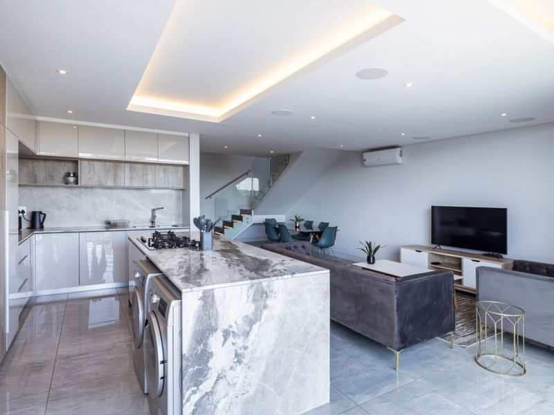 Modern open-plan living area featuring a sleek kitchen with marble island, gas hob, integrated appliances, and a contemporary staircase with glass balustrade.