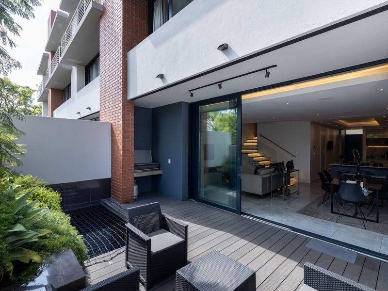 Modern covered patio featuring timber decking, a built-in braai, and a water feature, seamlessly connecting to the open-plan interior living space via sliding doors.