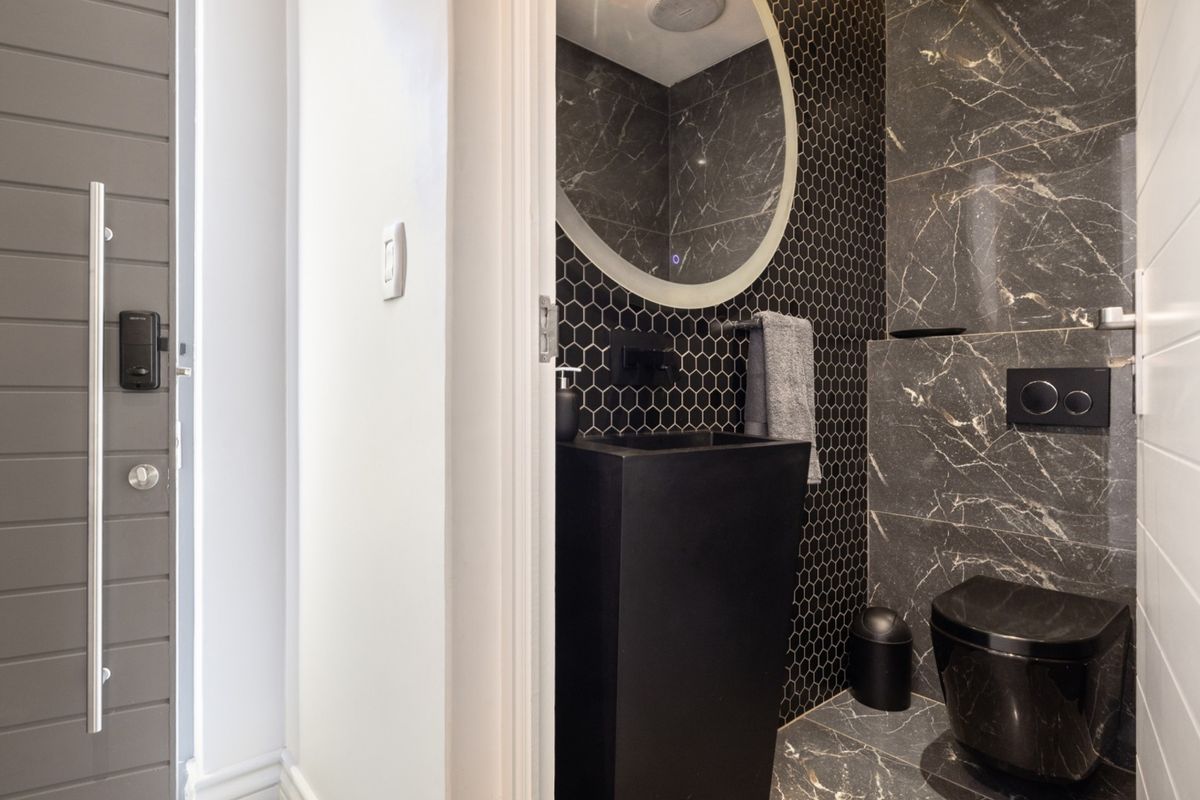 Modern guest bathroom featuring dark marble-look tiling, a sleek black pedestal basin, wall-mounted toilet with concealed cistern, and a contemporary backlit circular mirror.