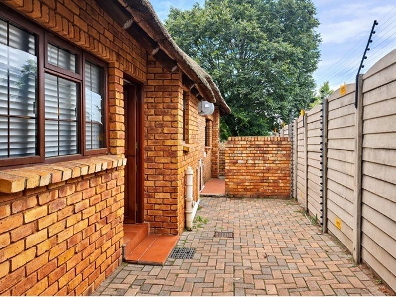 A side exterior view of the property featuring face brick walls, a thatched roof, paved walkway, and secure perimeter walling with electric fencing.
