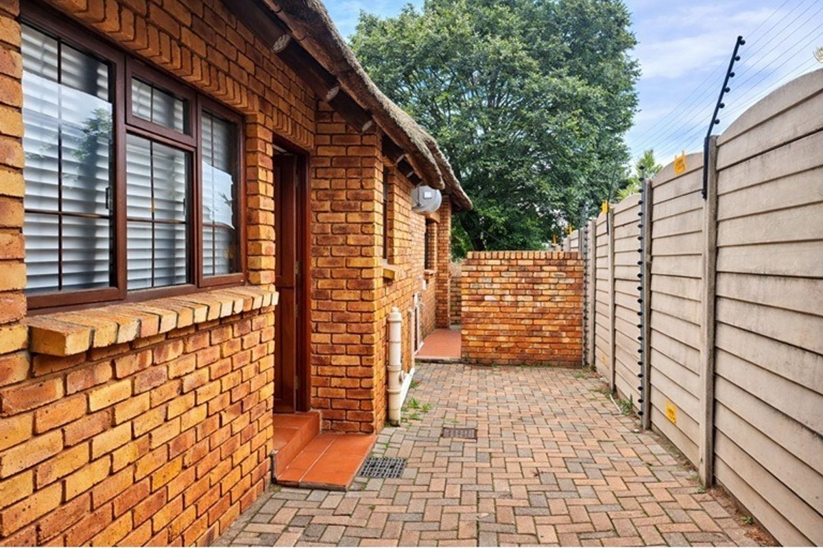 A side exterior view of the property featuring face brick walls, a thatched roof, paved walkway, and secure perimeter walling with electric fencing.
