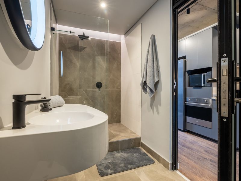 Modern bathroom featuring a wall-mounted vanity, black fixtures, and a walk-in shower with large format tiling, adjacent to a contemporary kitchen area.