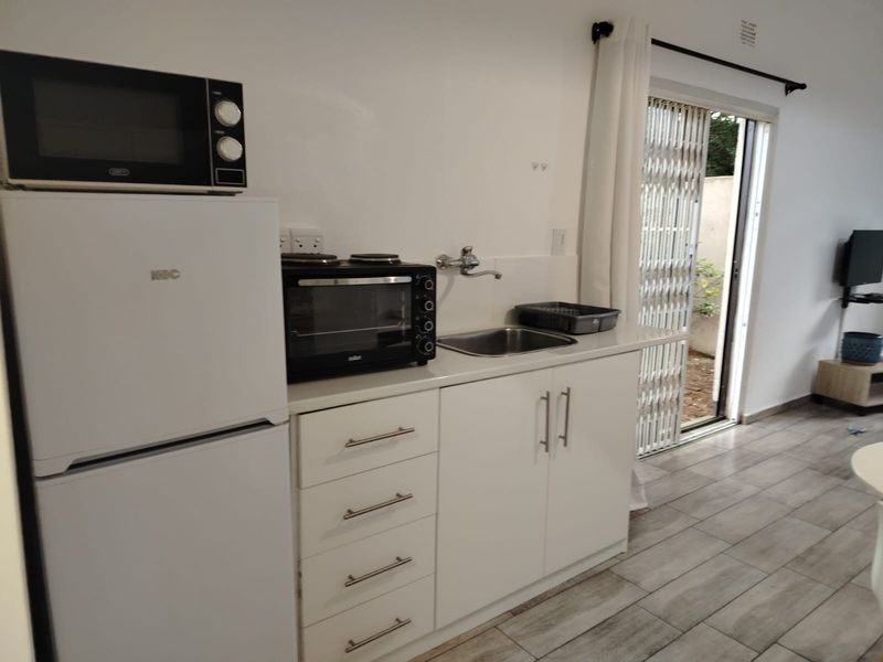 Compact kitchenette featuring white cabinetry, stainless steel sink, and tiled flooring, with direct access to the outdoor area through a sliding security door.