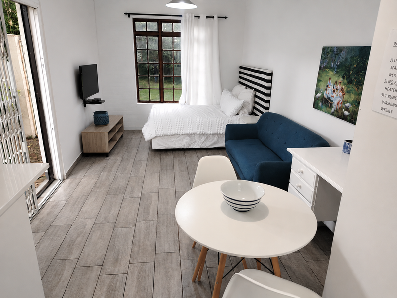 A bright open-plan studio cottage featuring modern tiled flooring, a timber-framed window, and direct access to an outdoor area via a security gate.
