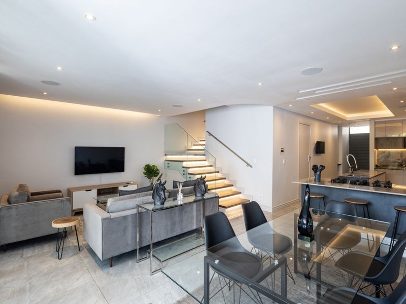 Modern open-plan living area featuring a sleek kitchen with stone island, integrated appliances, and a striking staircase with glass balustrade and ambient lighting.