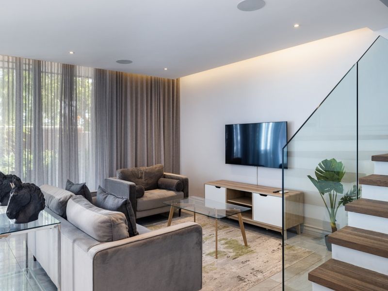 Modern living room featuring a glass balustrade staircase, recessed ceiling lighting, and large floor-to-ceiling windows with elegant drapery.