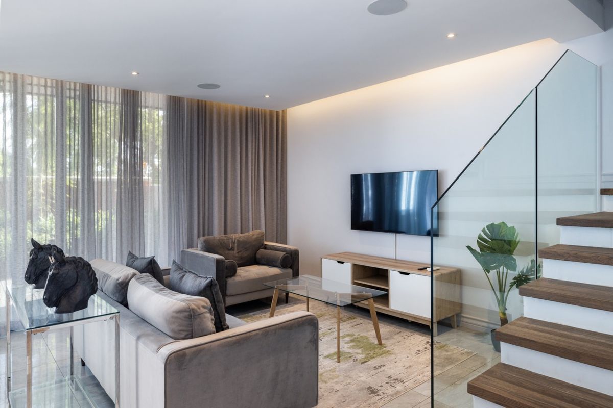 Modern living room featuring a glass balustrade staircase, recessed ceiling lighting, and large floor-to-ceiling windows with elegant drapery.