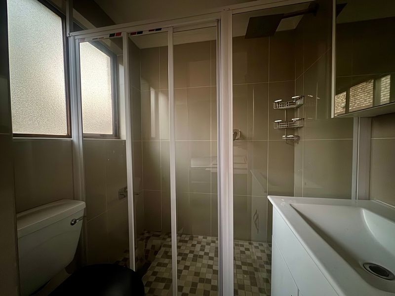 A functional bathroom featuring a shower enclosure, vanity basin, toilet, and neutral wall tiling with a frosted window for privacy.