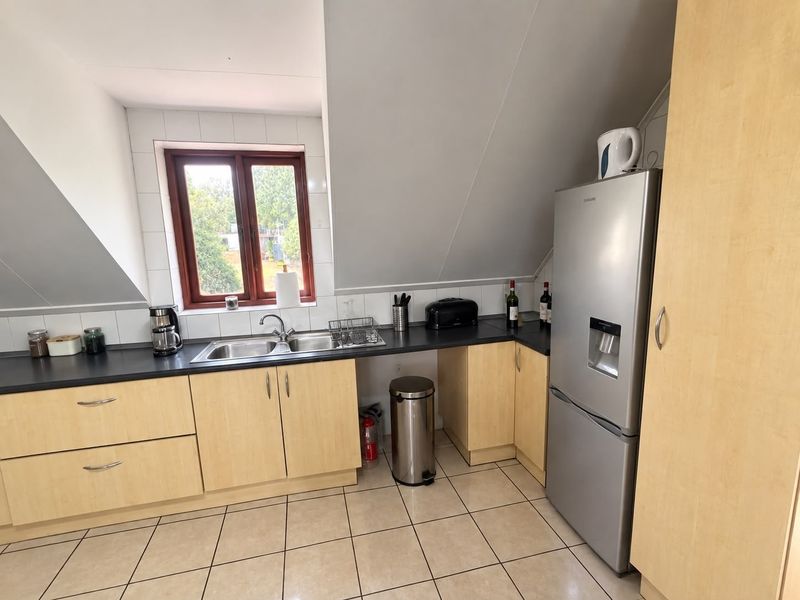 A functional kitchen featuring light wood-tone cabinetry, dark laminate countertops, tiled splashbacks, and ceramic floor tiles under a sloped ceiling.